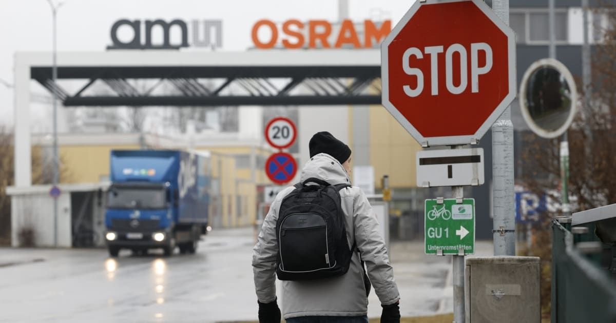 A man wearing a backpack walks past a STOP sign, near an OSRAM factory, with a blue truck driving on a wet road.