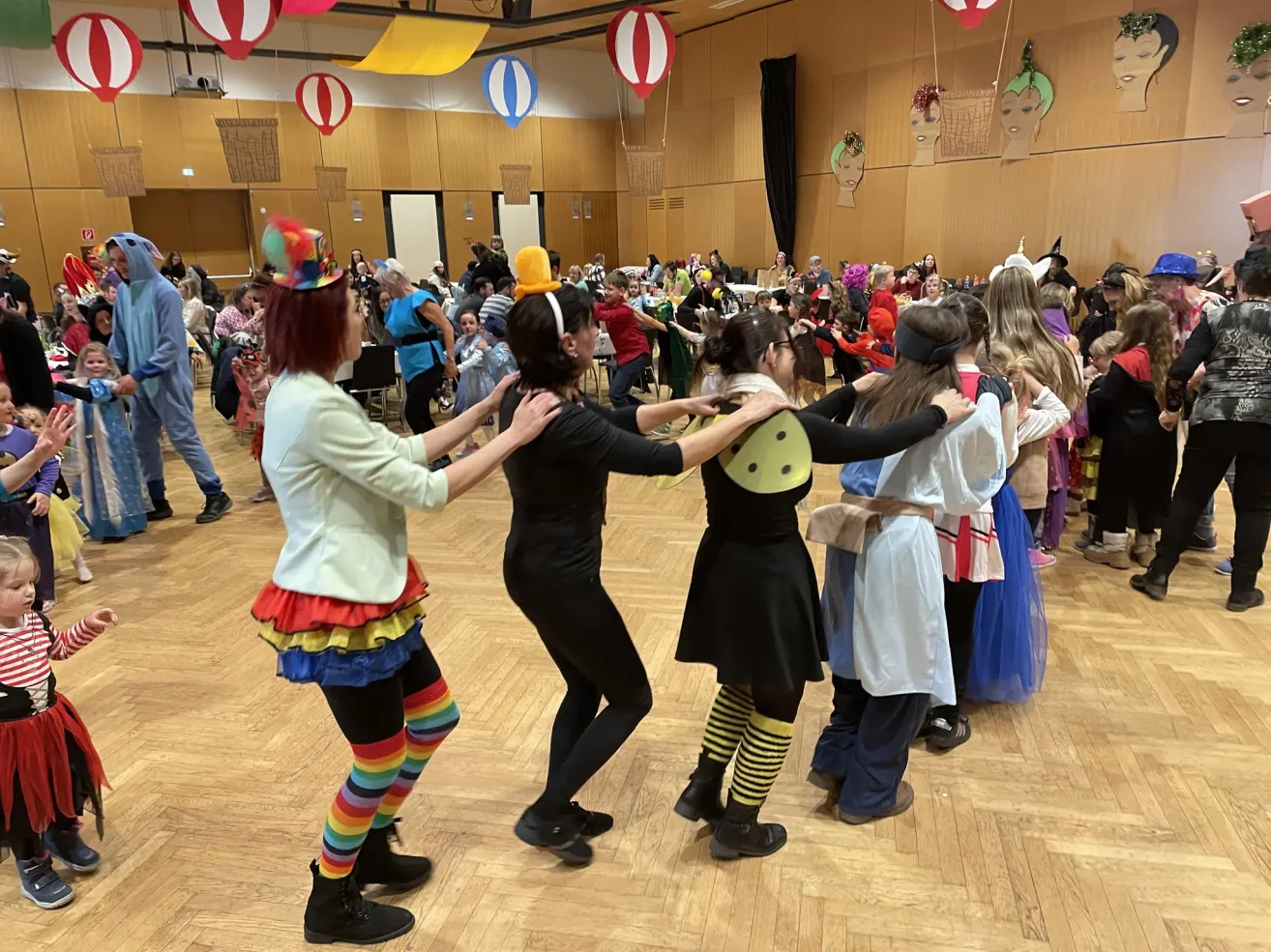 A group of people in costumes dance in a large hall, forming a circle with others behind them. Colorful balloons hang from the ceiling.