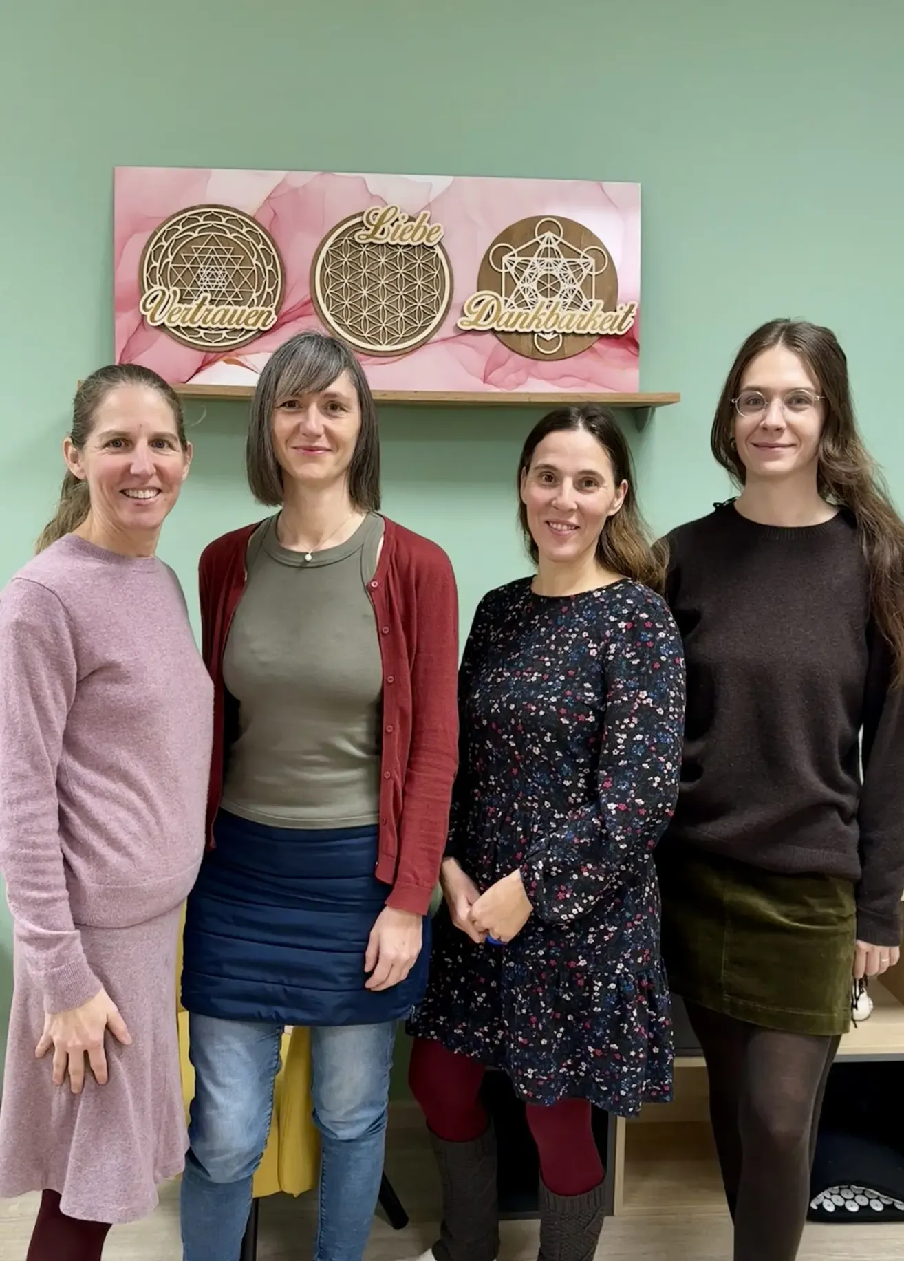 Four women stand together in front of a green wall, smiling for a photo. They wear different colored clothes. A sign with 'Liebe,' 'Vertrauen,' and 'Dankbarkeit' is mounted above them.