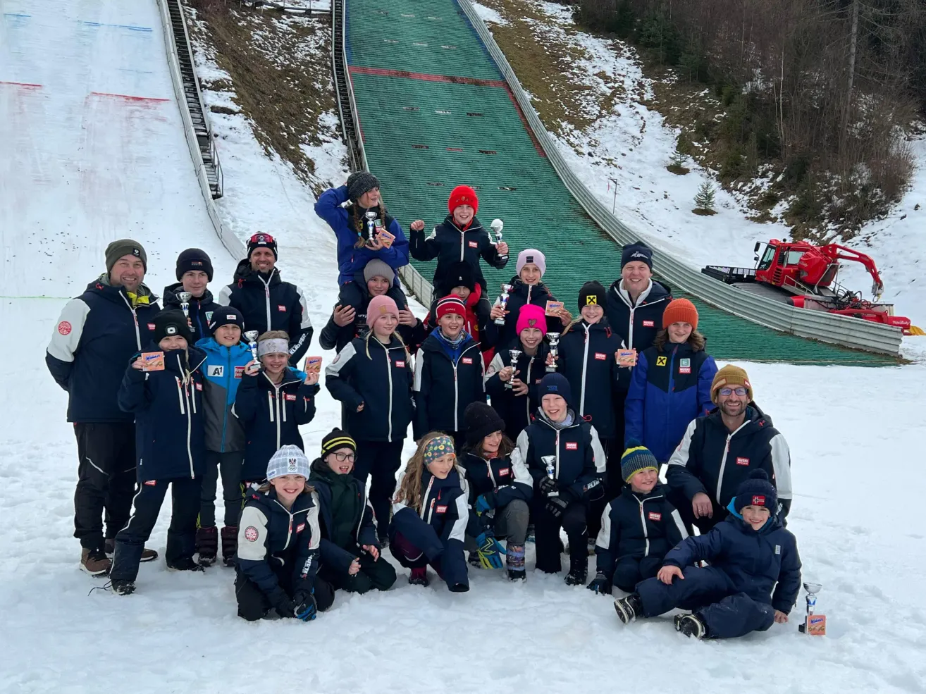 A group of people in winter clothing pose for a photo in the snow. Some hold trophies. They are in front of a ski jump with a green slope.