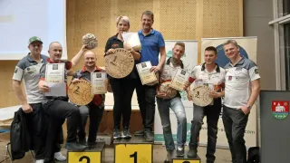 Seven individuals stand on a stage, three holding trophies, wearing sneakers, and holding certificates. They are smiling and posing for a photo. The stage has a podium with numbers 1, 2, and 3.