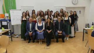 A group of people dressed in traditional attire pose for a photograph in a room with wooden floors, a white wall, and a clock. Some are seated while others stand behind them.