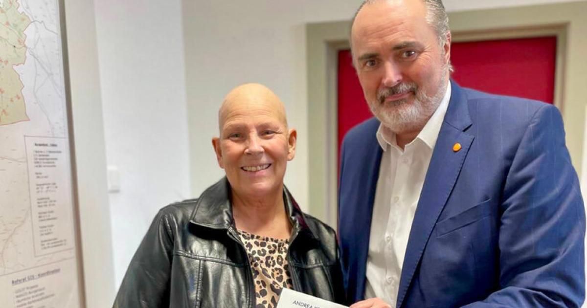 A bald woman and a bearded man are standing close together, smiling, and posing for a photo. The woman is holding a piece of paper, while the man is wearing a blue suit.