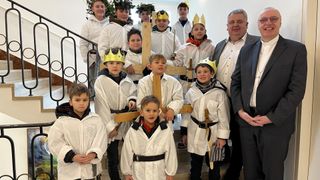 A group of children dressed in white, wearing crowns, and holding wooden crosses pose for a photo on stairs with a man in a suit smiling.