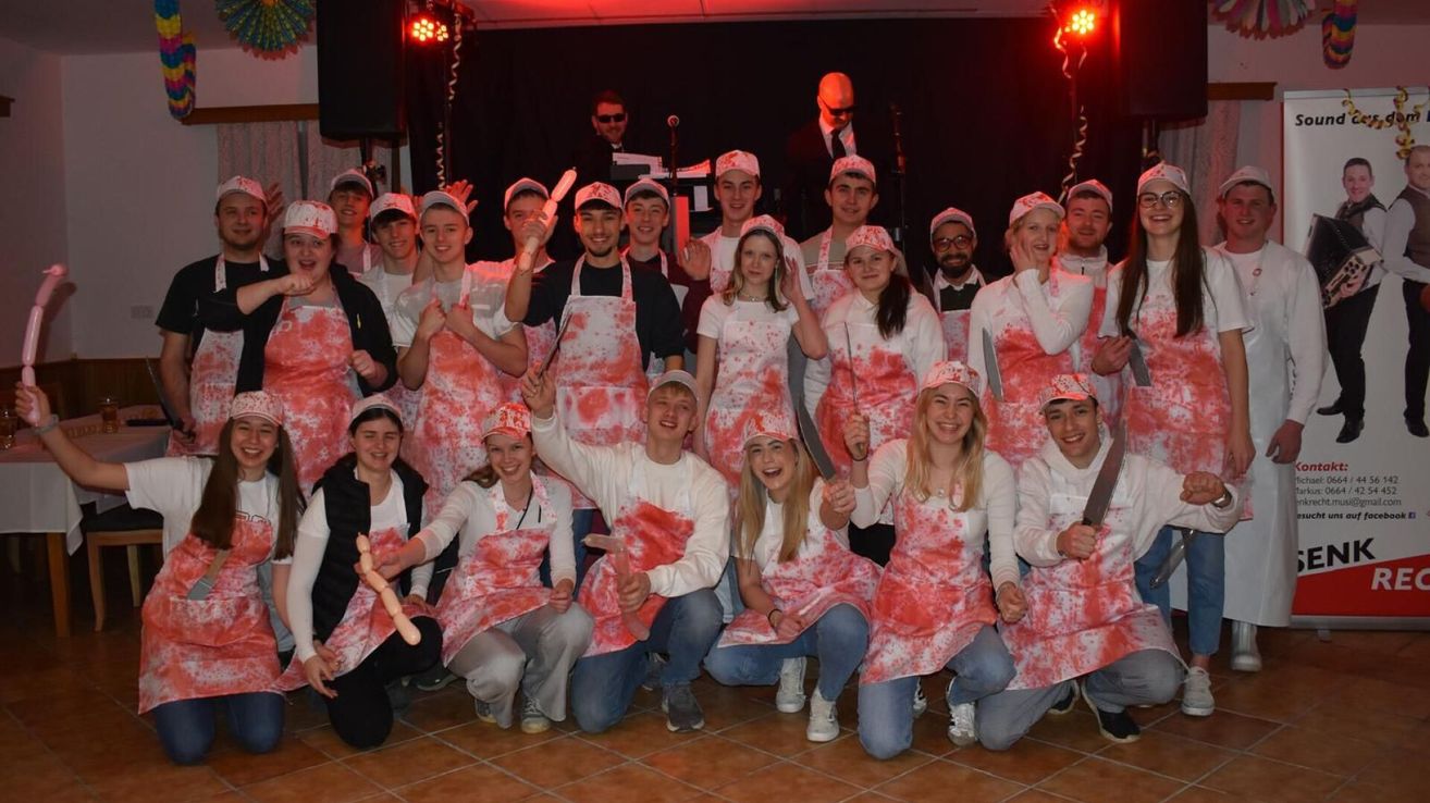 A group of people dressed in red and white aprons, holding knives and sausages, are posing for a photo in a room with red lights. Behind them, two people are standing, one wearing glasses and the other with a microphone.