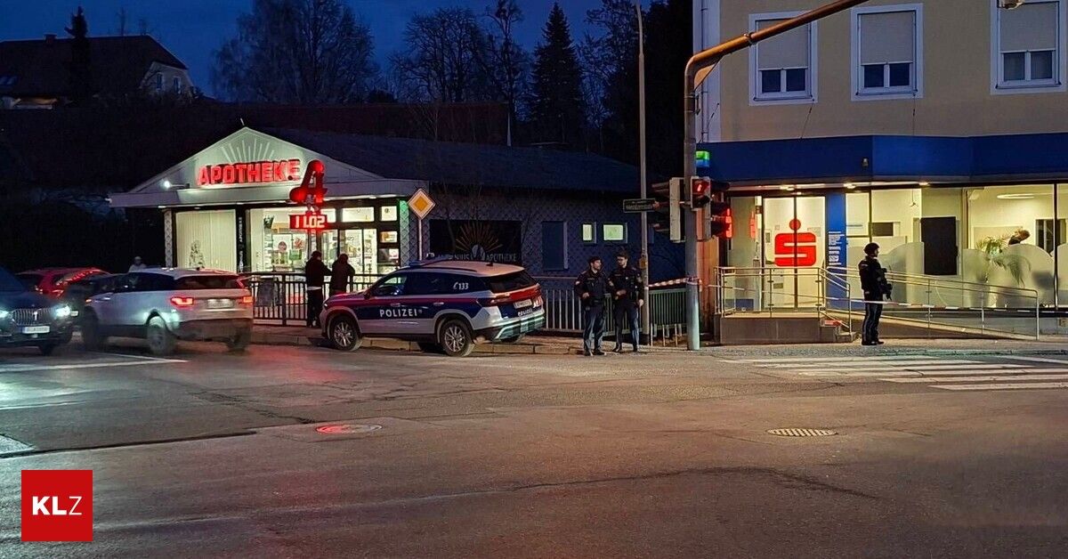 In der Nacht stehen zwei Polizeibeamte neben einem Polizei-SUV vor einer Apotheke, mit zwei Fußgängern am Eingang.