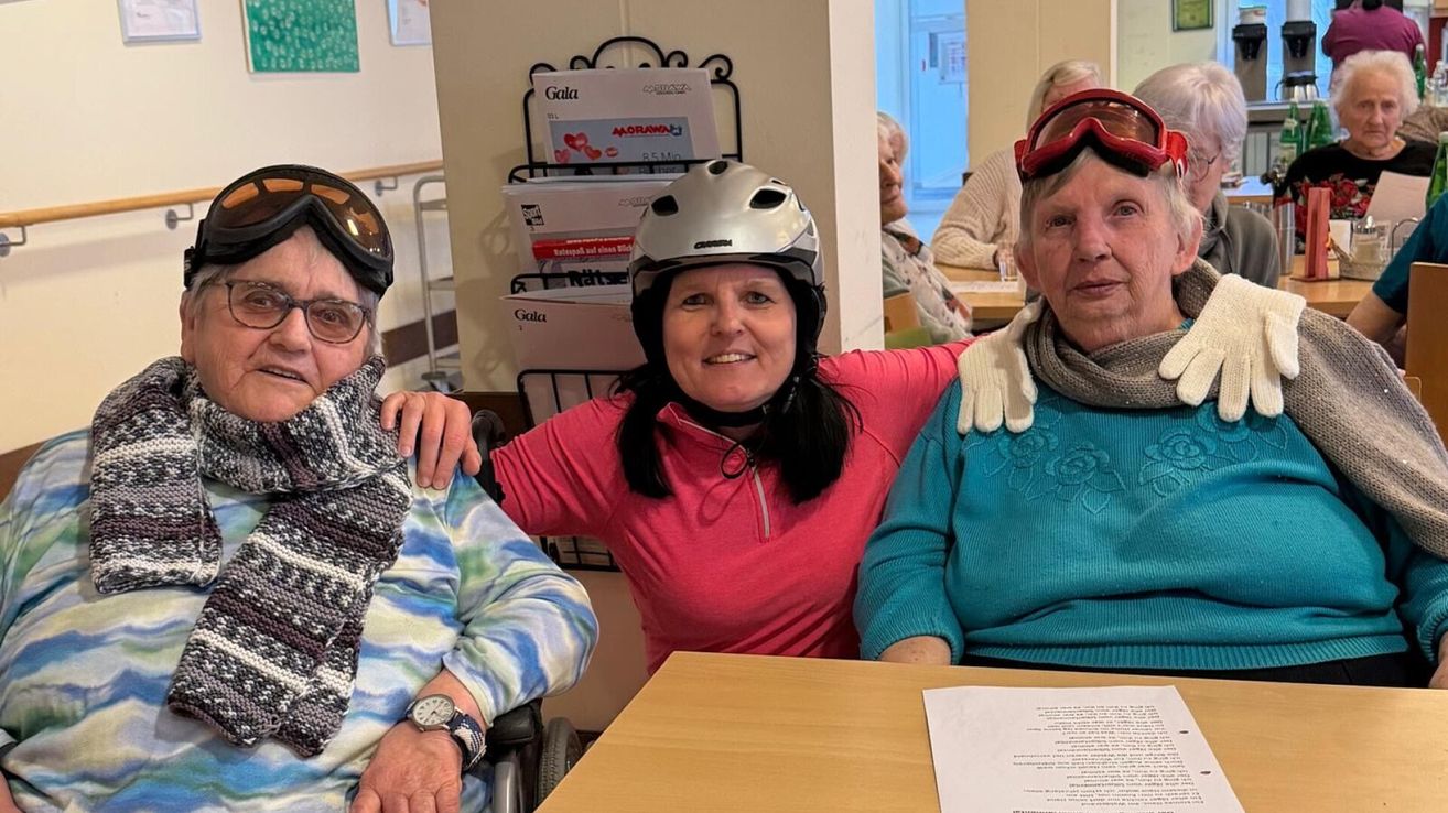 Three women in a room. One wearing a helmet and smiling, another wearing goggles, and the third wearing a scarf. A table with papers is in front of them.