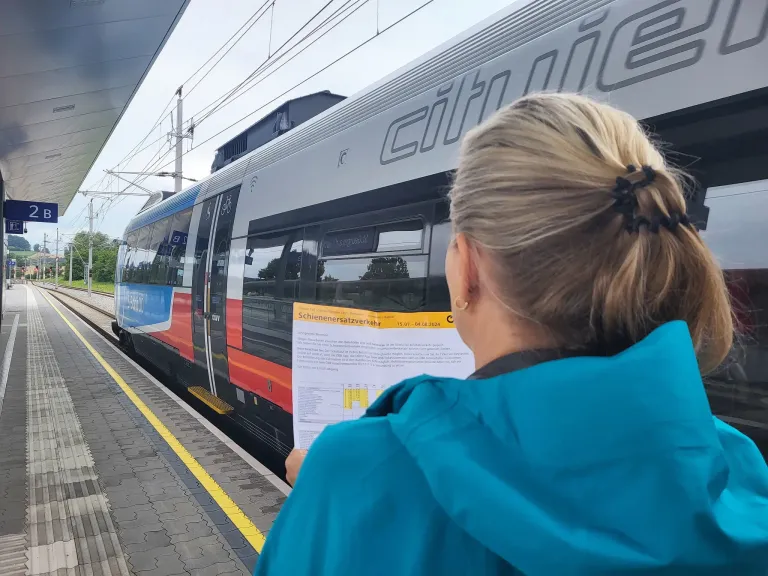 A woman reads a paper at a train station. The train reads 'citie' and has red and white windows. The woman wears a blue jacket.