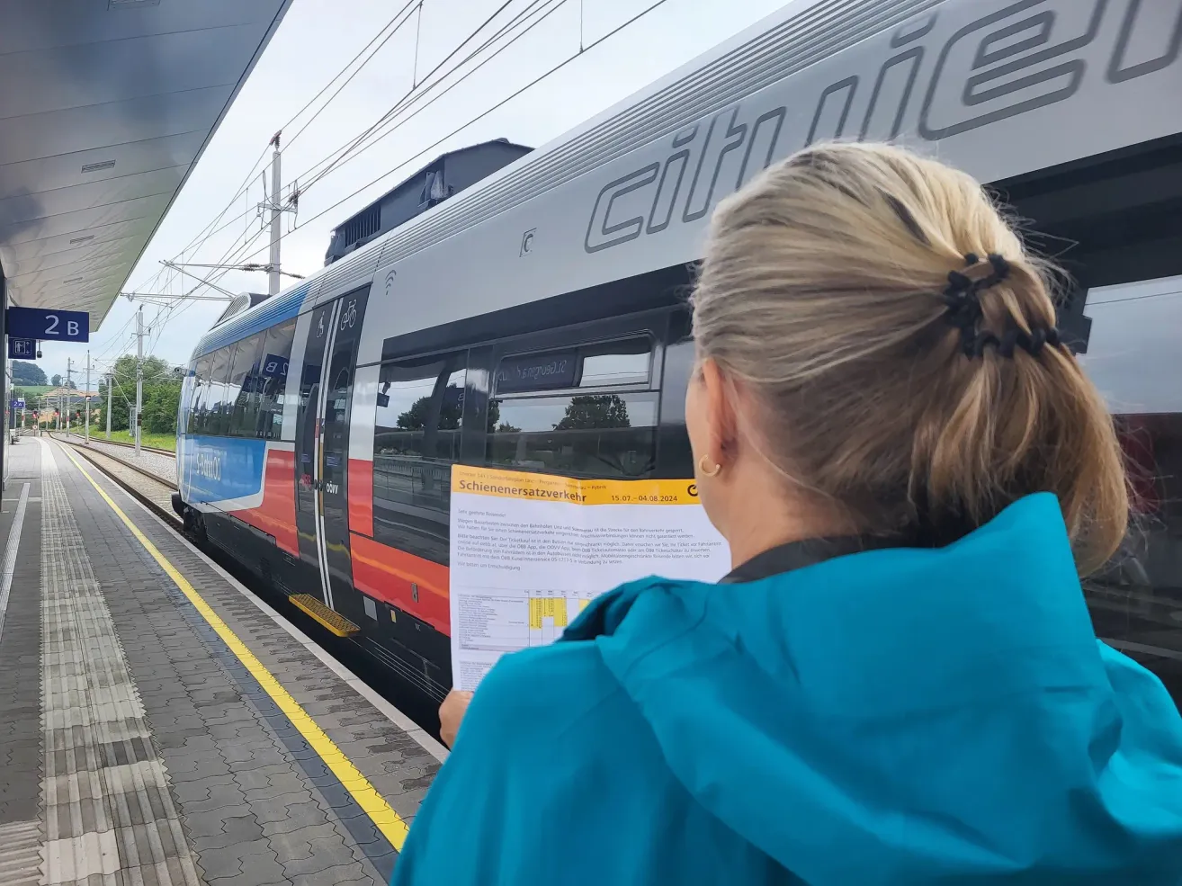 A woman reads a paper at a train station. The train reads 'citie' and has red and white windows. The woman wears a blue jacket.