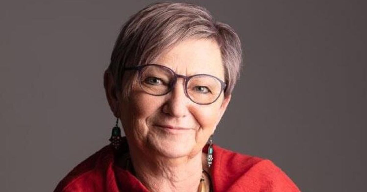 A woman with short gray hair wearing glasses and a red top smiles at the camera. She has earrings with green and turquoise stones.