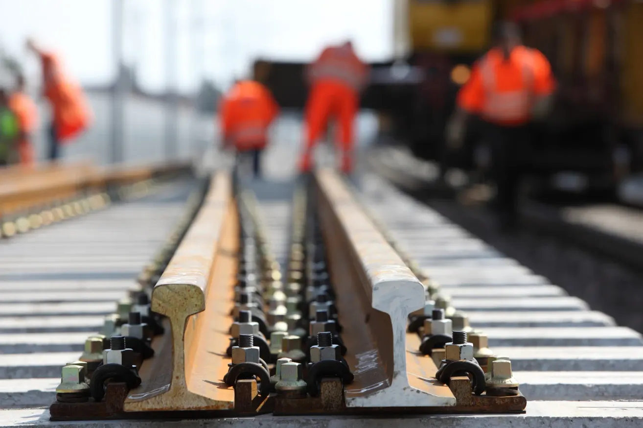 A blurred background with three workers in orange safety vests and black pants working on a railway track with steel rails and bolts.