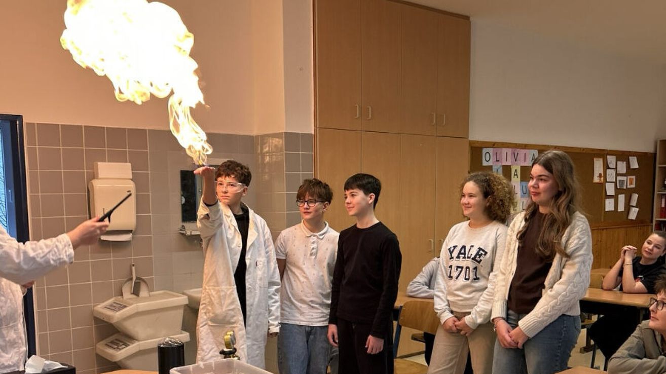In a classroom, a student is holding a flame while another student is raising his hand. A few other students are standing behind them. Behind them is a sink and a cabinet.