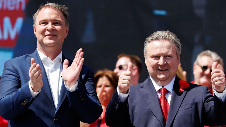 Two men in suits clapping and giving thumbs up. One man has gray hair, the other has dark hair. In the background, a woman in red waves.