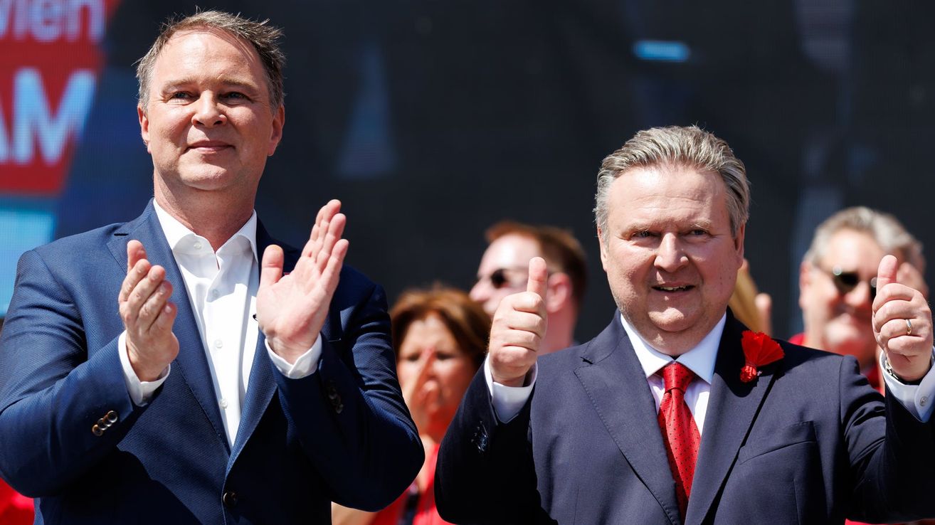 Two men in suits clapping and giving thumbs up. One man has gray hair, the other has dark hair. In the background, a woman in red waves.