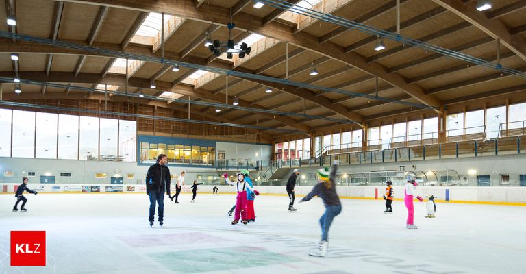 Several people are ice skating in an indoor ice rink. The rink has a wooden ceiling with lights and glass windows. Some are wearing winter clothes, while others are in casual attire.