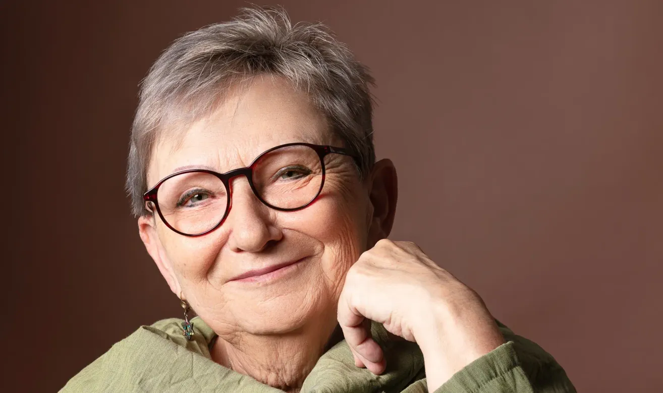 An older woman with gray hair, wearing glasses, and a green top. She is smiling and posing for a picture. She has a light brown skin tone.