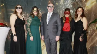 Five people are standing close together, wearing masks and black or colored dresses, posing for a photo in front of a patterned backdrop.