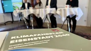 A group of people seated at a table with a book titled Klimafahrplan Eisenstadt 2040. A monitor is behind them.