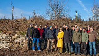 A group of people dressed in winter attire are posing for a photo in a muddy area with a stone wall and barren trees in the background.