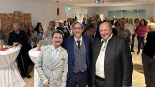 Three men in formal attire stand close together, smiling for a photo at an event, with a crowd of people and tables in the background.