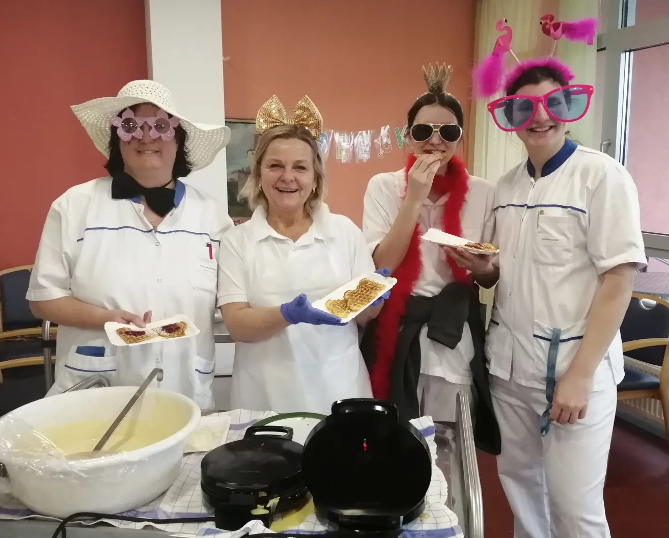 Four individuals dressed in white uniforms are posing with waffles in a kitchen setting. One person is wearing a crown, while another holds a red scarf. A waffle maker is on the table.