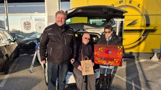 Three people are standing in front of a car with an open trunk. The man on the left is holding a bag. The woman on the right is holding a picture frame. The man in the middle is holding a cigarette.