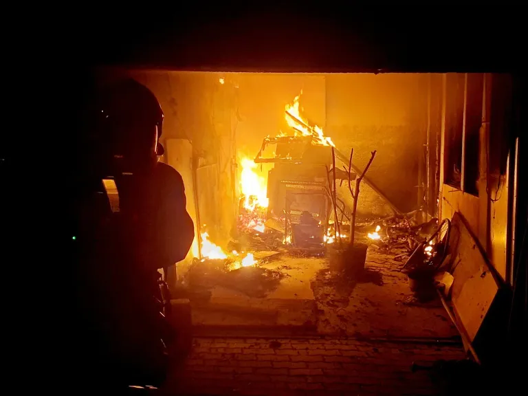 A firefighter in a helmet stands in a garage with a burning vehicle. The flames illuminate the space, casting shadows on the walls.