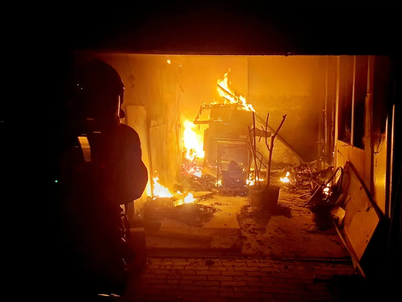 A firefighter in a helmet stands in a garage with a burning vehicle. The flames illuminate the space, casting shadows on the walls.