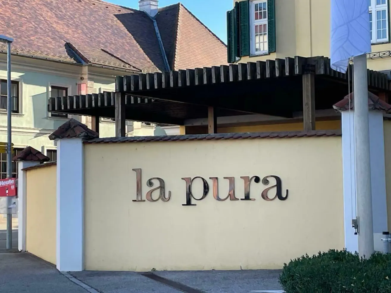 A building with a brown roof has a white wall with the word la pura in metal letters. It is surrounded by a stone wall with a black awning above.