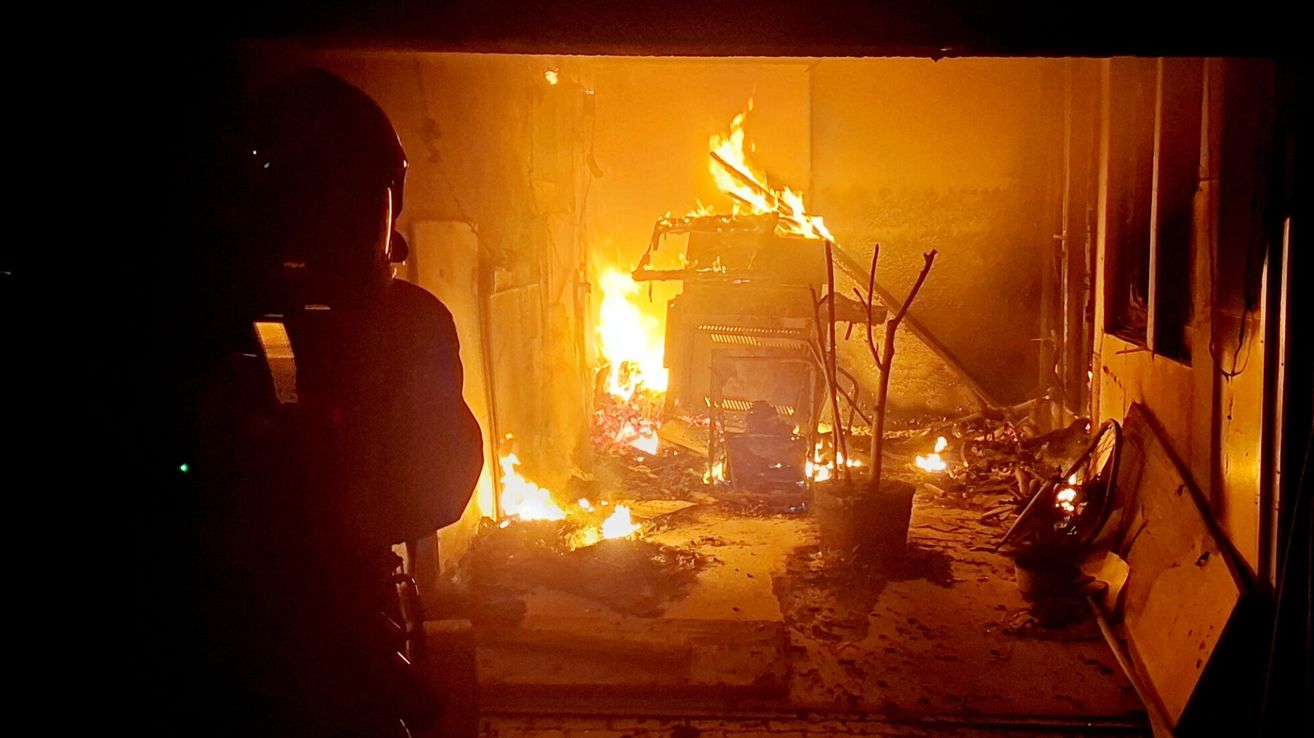 A firefighter stands in a room with a fire, observing the damage. The fire has burned furniture and the walls.