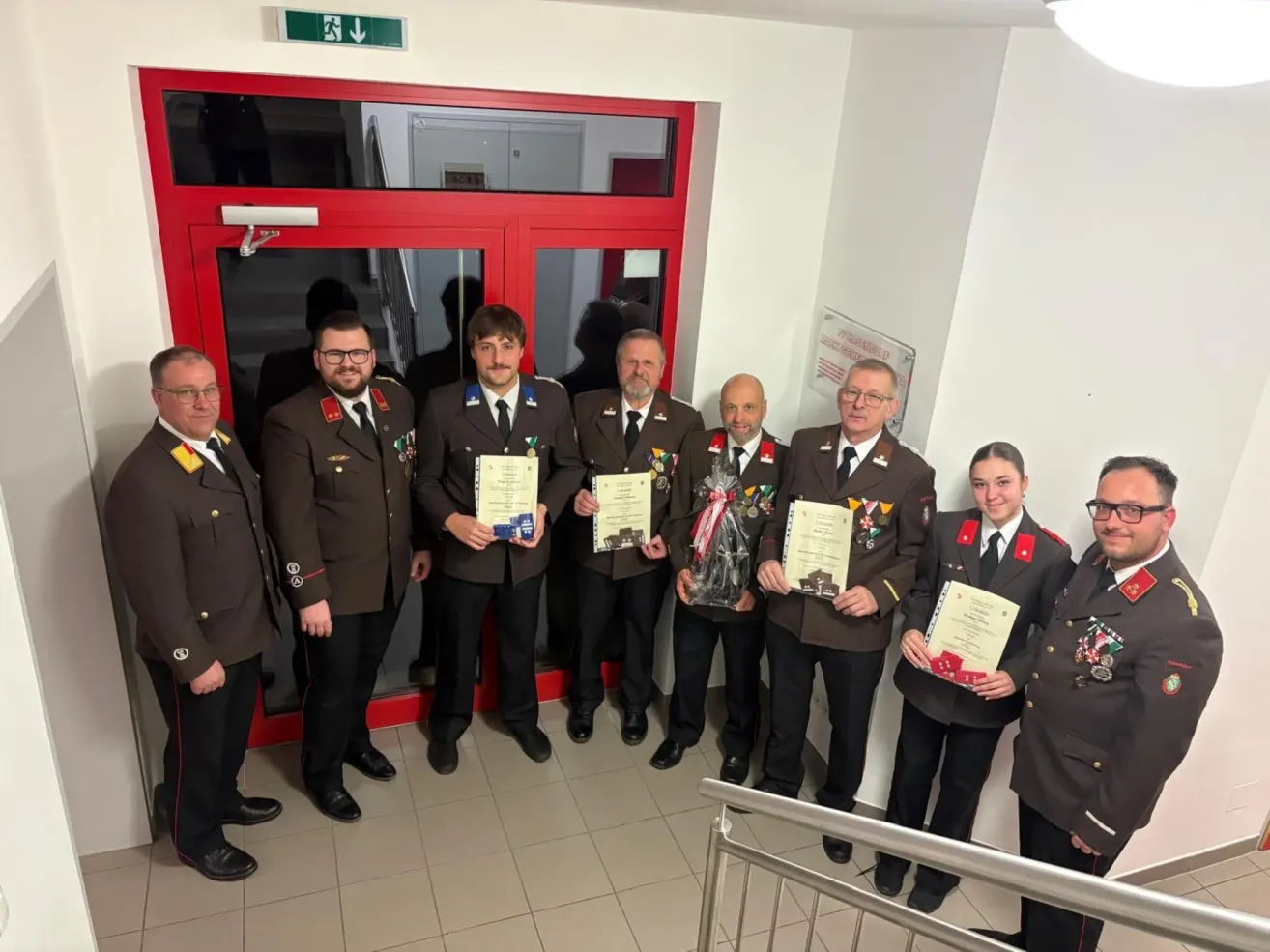 A group of uniformed individuals stand in front of a red door, holding certificates and awards, likely celebrating a significant achievement.