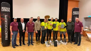 Group photo of winners in yellow shirts holding trophies and medals, with a large trophy in the center, standing on a wooden floor.