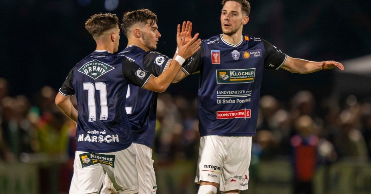 Three soccer players celebrate a goal, wearing blue jerseys with sponsors' logos. The player in the middle, with his arms raised, seems to be shouting. The player on the left wears number 71. The background shows a blurred crowd.