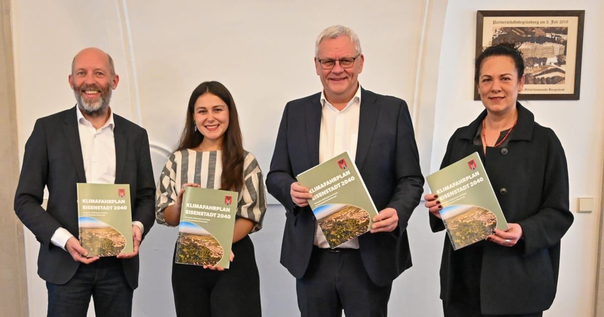 Four people stand together holding up books with 'Klimafahrplan Eisenstadt 2040' on the cover. They are in a light room with white walls.