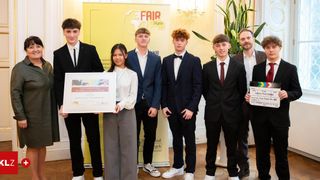 A group of young individuals, dressed formally, are posing for a photo. A woman holds a certificate, and a man next to her holds a clapperboard. Behind them is a banner with the word 'FAIR'.