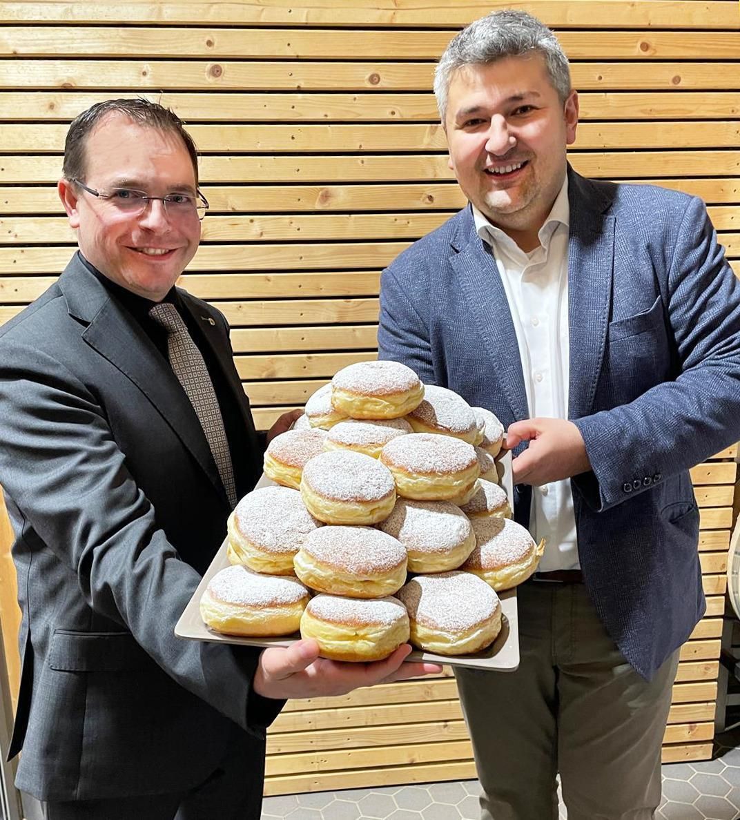 Two men in business attire are holding a tray of powdered donuts, smiling in front of a wooden wall.