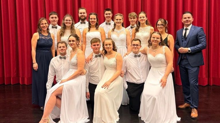 Eine Gruppe junger Erwachsener, sowohl Männer als auch Frauen, sind in weißen Kleidern und Fliegen gebunden und posieren für ein Foto vor einem roten Vorhang.