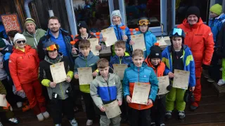 A group of young boys and girls are standing together, holding certificates and posing for a photograph. They are wearing winter outfits and some have helmets and goggles. They are standing on a wooden floor in front of a glass wall.