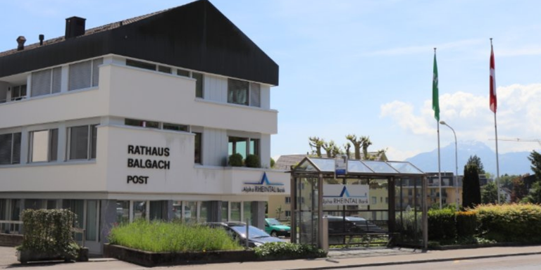 The Rathaus Balgach Post building has a flag and a signboard. A bus stop with a bench is in front. A car is parked nearby. Trees and mountains are in the distance.