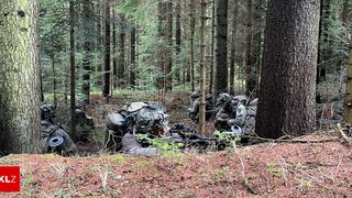 A group of soldiers equipped with camouflage uniforms and helmets are lying on the ground in a forest, using their rifles to aim at a target.