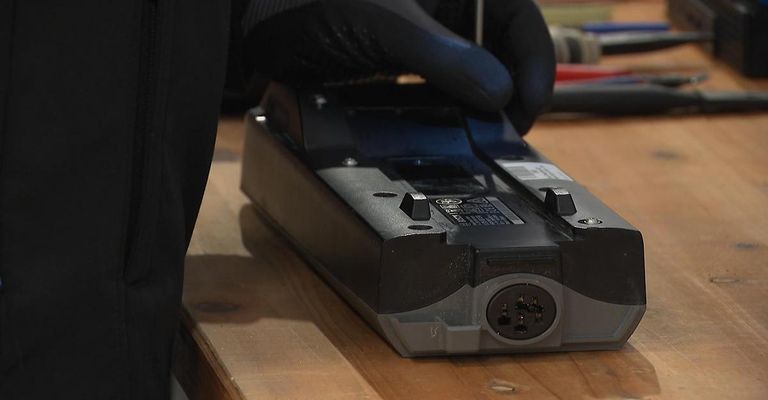 A person wearing black gloves is holding a black power supply with a plug on a wooden table.