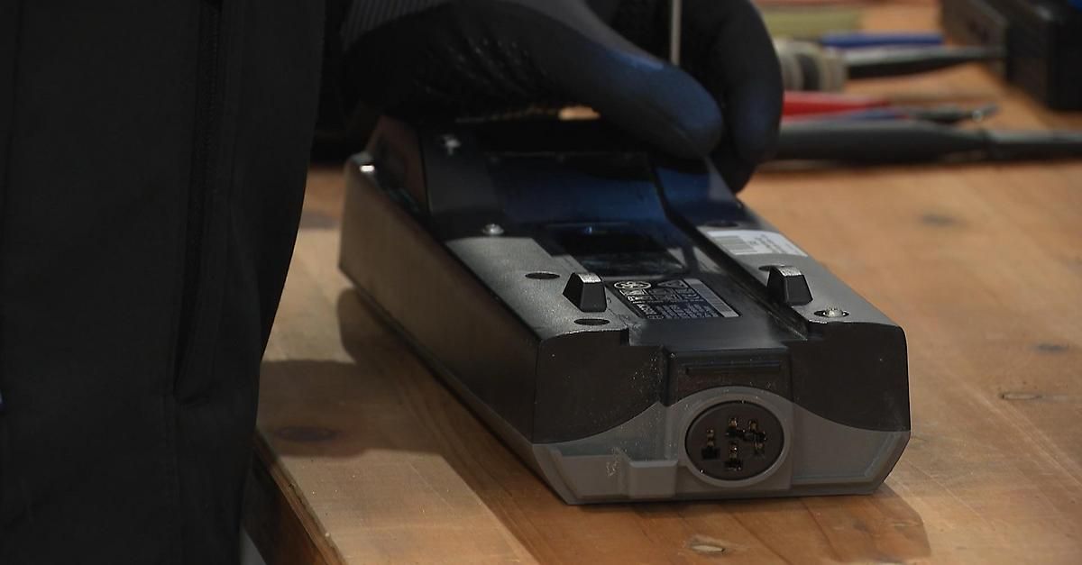 A person wearing black gloves is holding a black power supply with a plug on a wooden table.