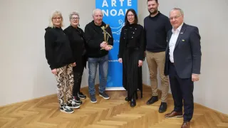 A group of individuals stands indoors in front of a blue banner with 'ARTE NOAL' written on it. The man in the middle holds a trophy.