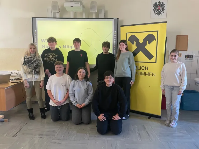 A group of students in a classroom with a yellow screen and a yellow banner reading 'Zlich Kommen'. The students are posing for a photo.