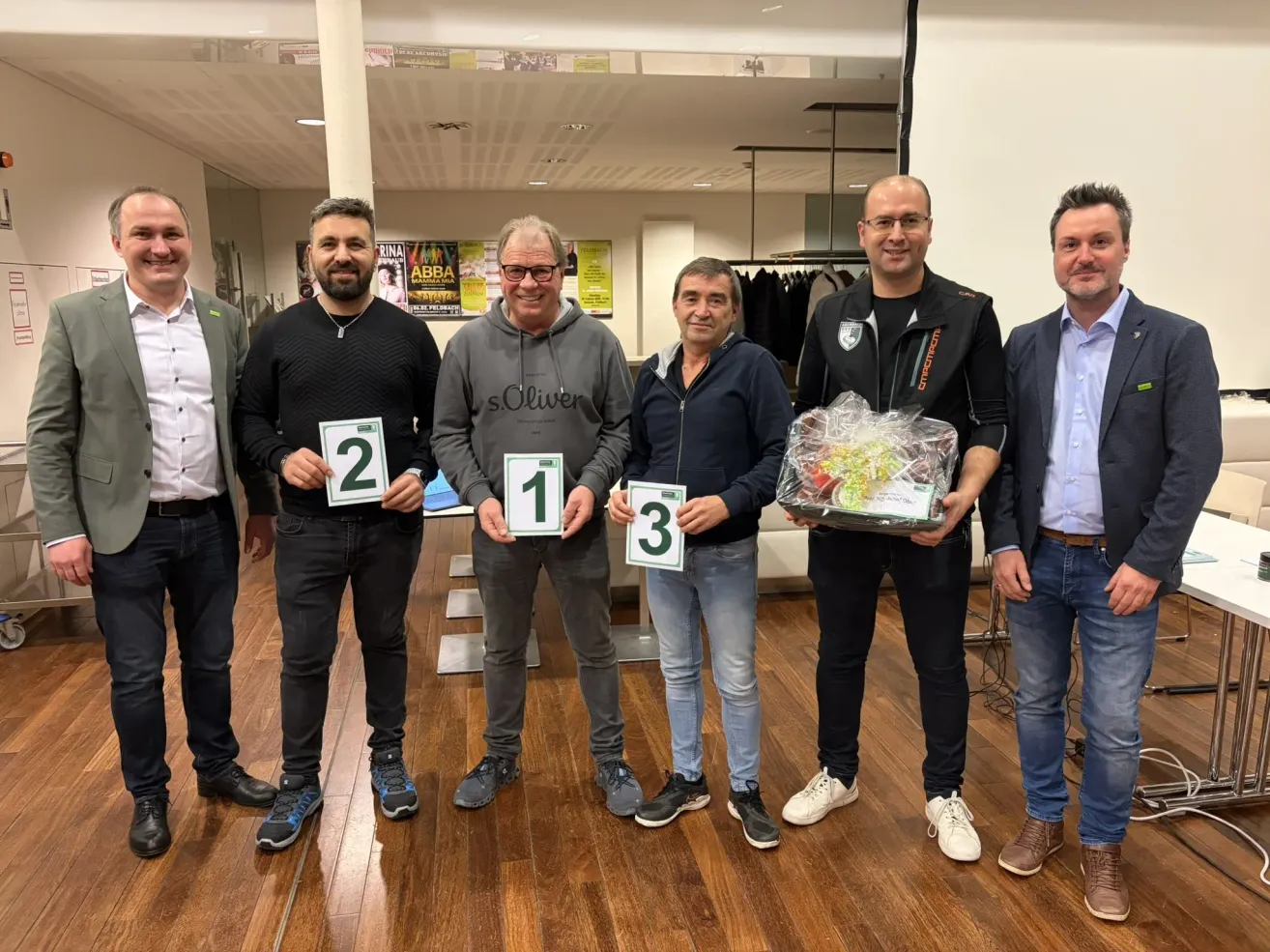 Five men stand together on a wooden floor, posing for a photo. Three of them hold numbers, one holds a gift basket, and one stands behind the man with the basket.