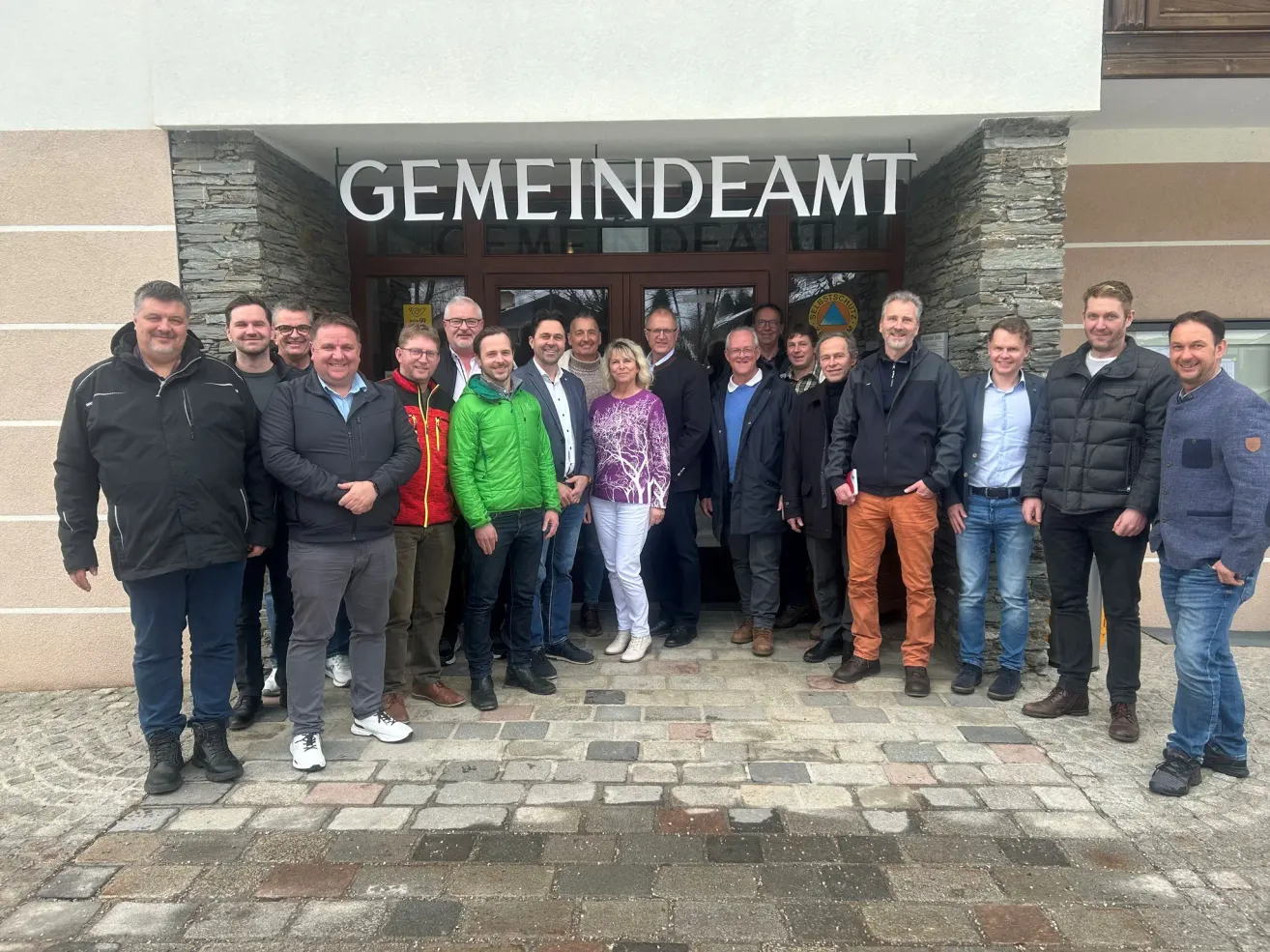 Eine Gruppe von Menschen in Winterkleidung steht vor einem Gebäude mit dem Schild 'GEMEINDEAMT' über ihnen. Sie posieren für ein Foto.