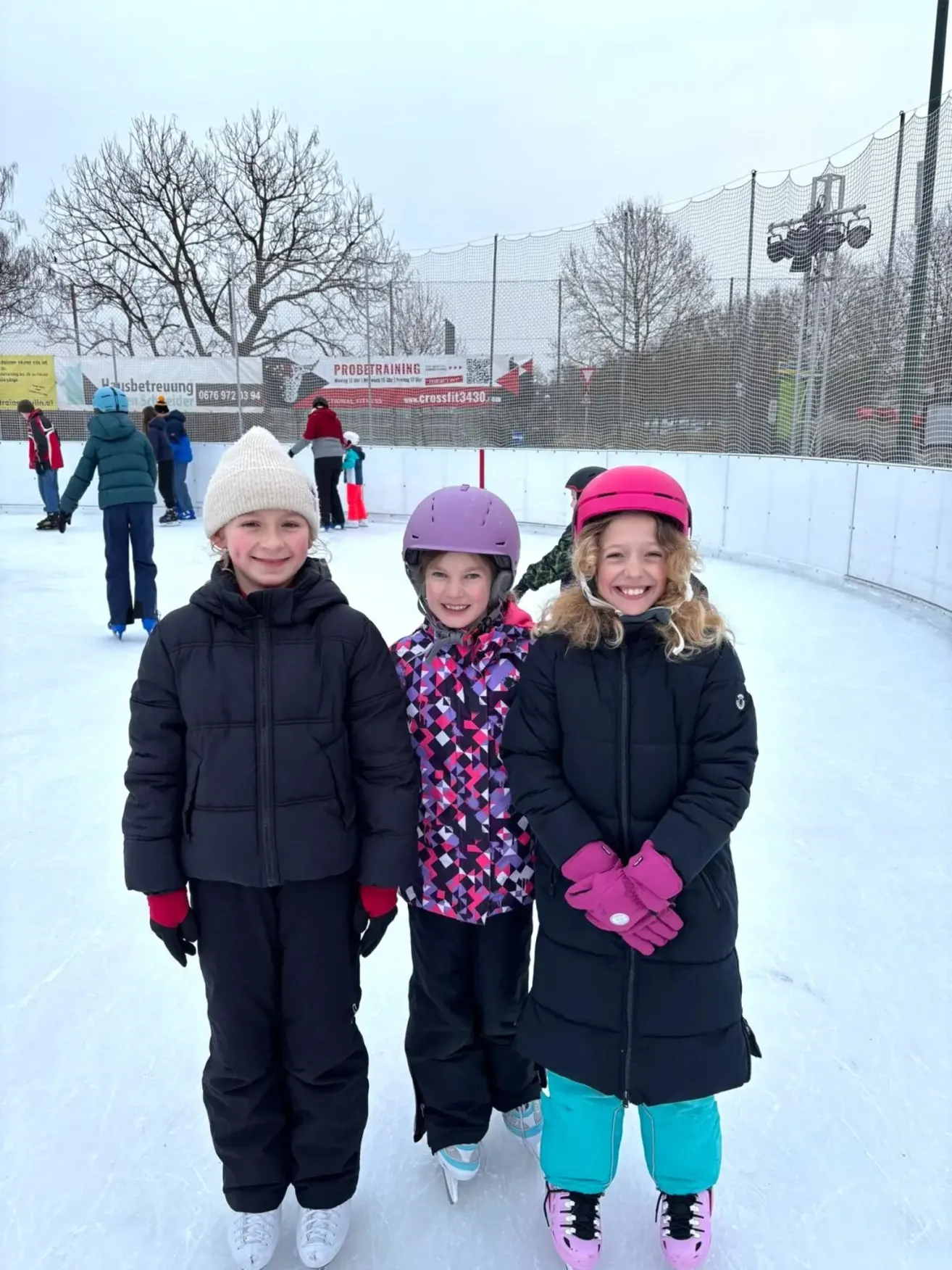 Three children wearing winter gear and helmets stand on an ice rink. A fence and trees are behind them.