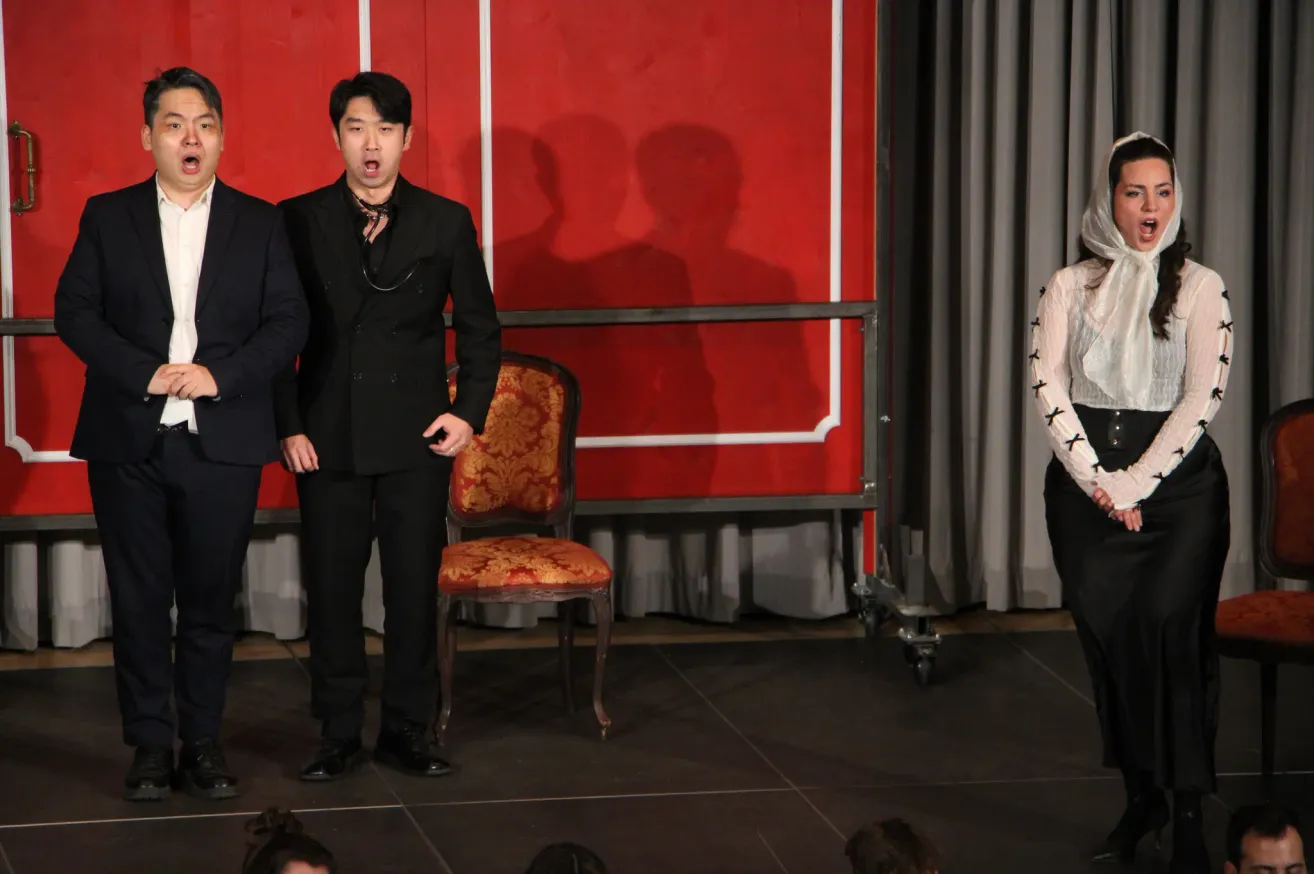 Two men and a woman stand on a stage. The man in the center wears a black suit and a scarf. An ornate chair is behind them, with a red backdrop and shadows on the wall.