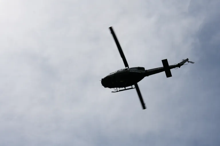 A black helicopter is flying in the sky with clouds. It has a long tail and two propellers. The sky is blue with white clouds.