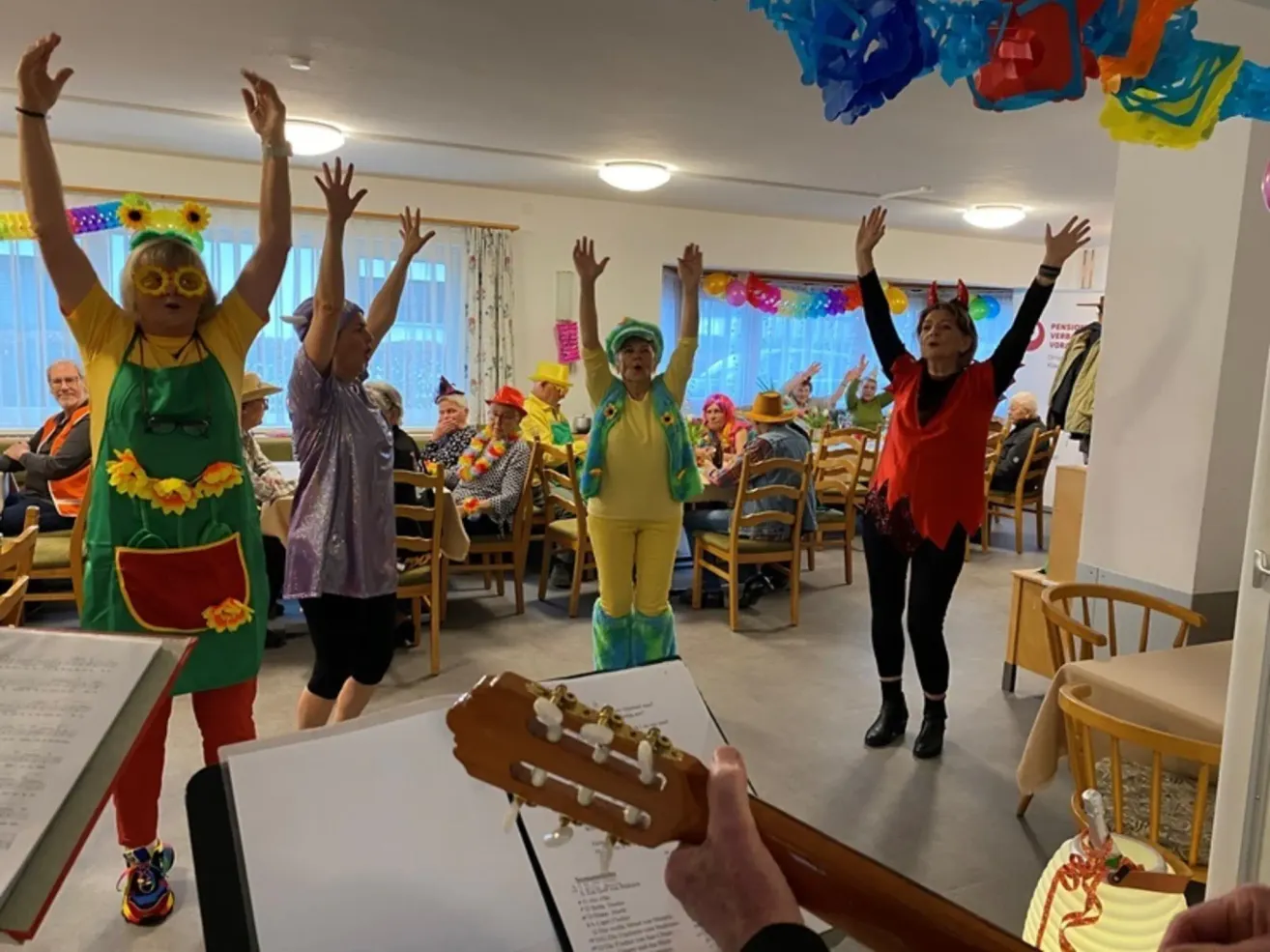 A group of elderly people are dancing in a room, with one person playing a guitar. Colorful decorations hang from the ceiling.